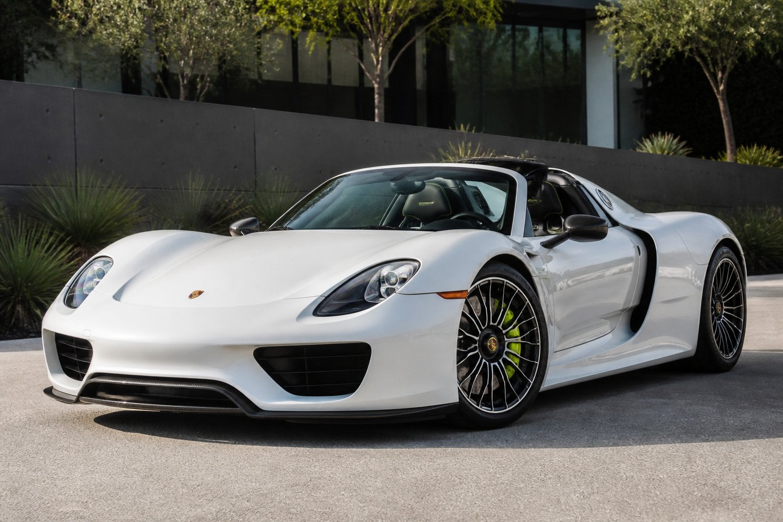 A white Porsche 918 Spyder parked outdoors with a modern city background.