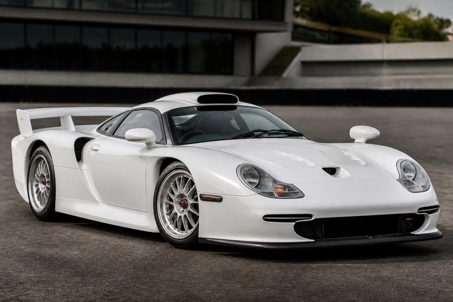 A white Porsche 911 GT1 Straßenversion sports car parked outdoors with a blurred racetrack background.