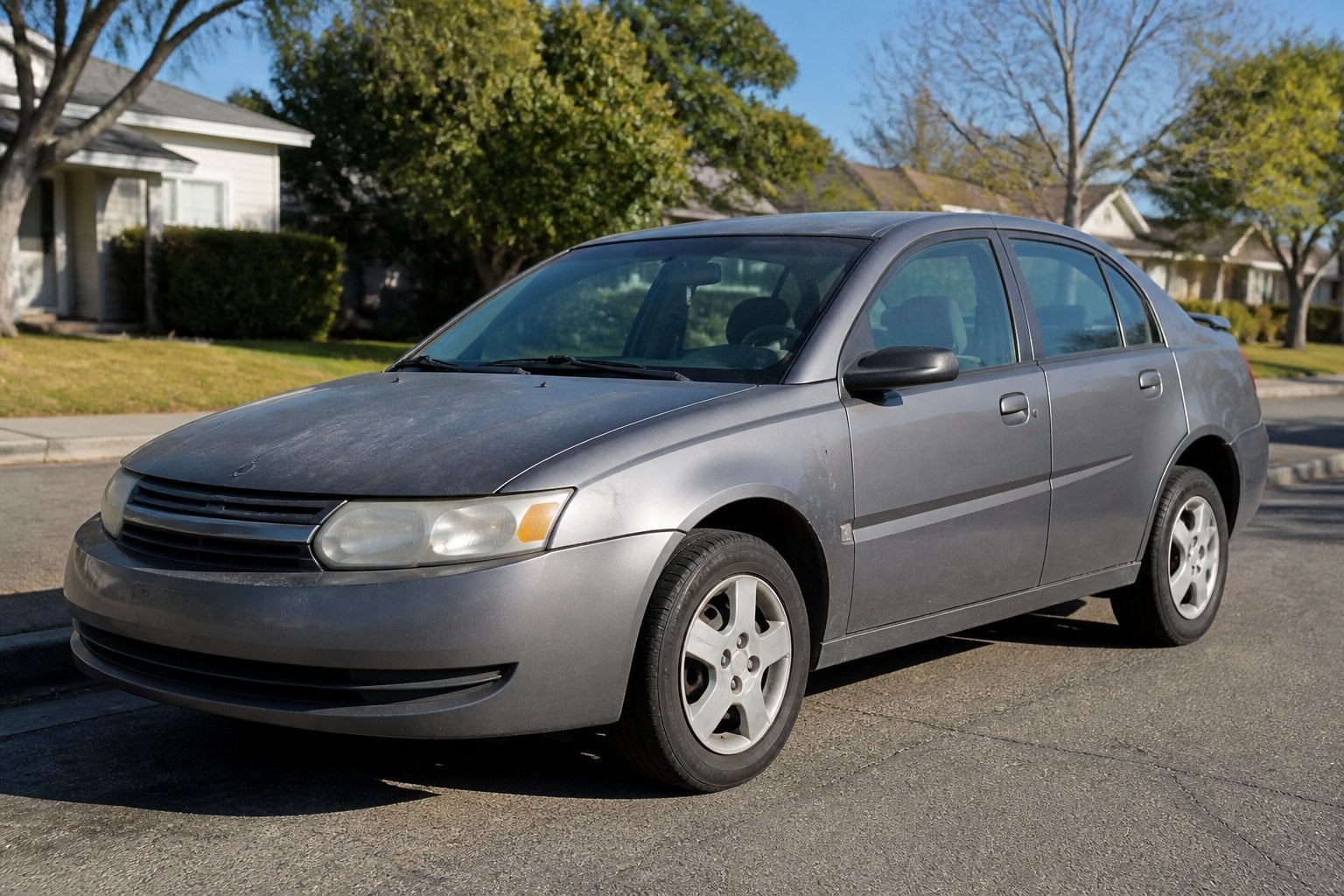 A worn 2003-2007 Saturn Ion parked on a suburban street with houses and trees in the background.