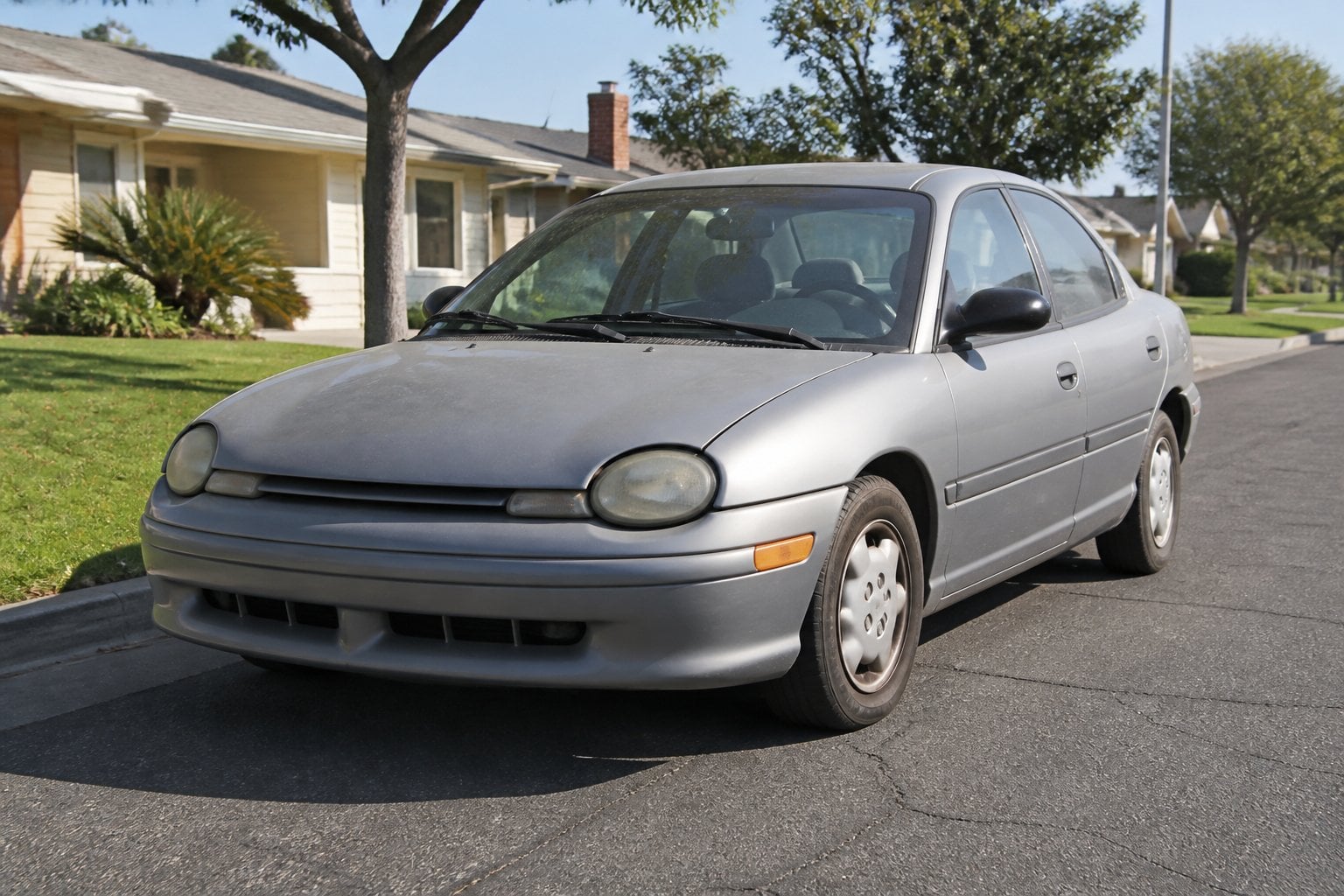 A worn Dodge Neon car parked on a suburban street with houses and trees in the background during daytime.