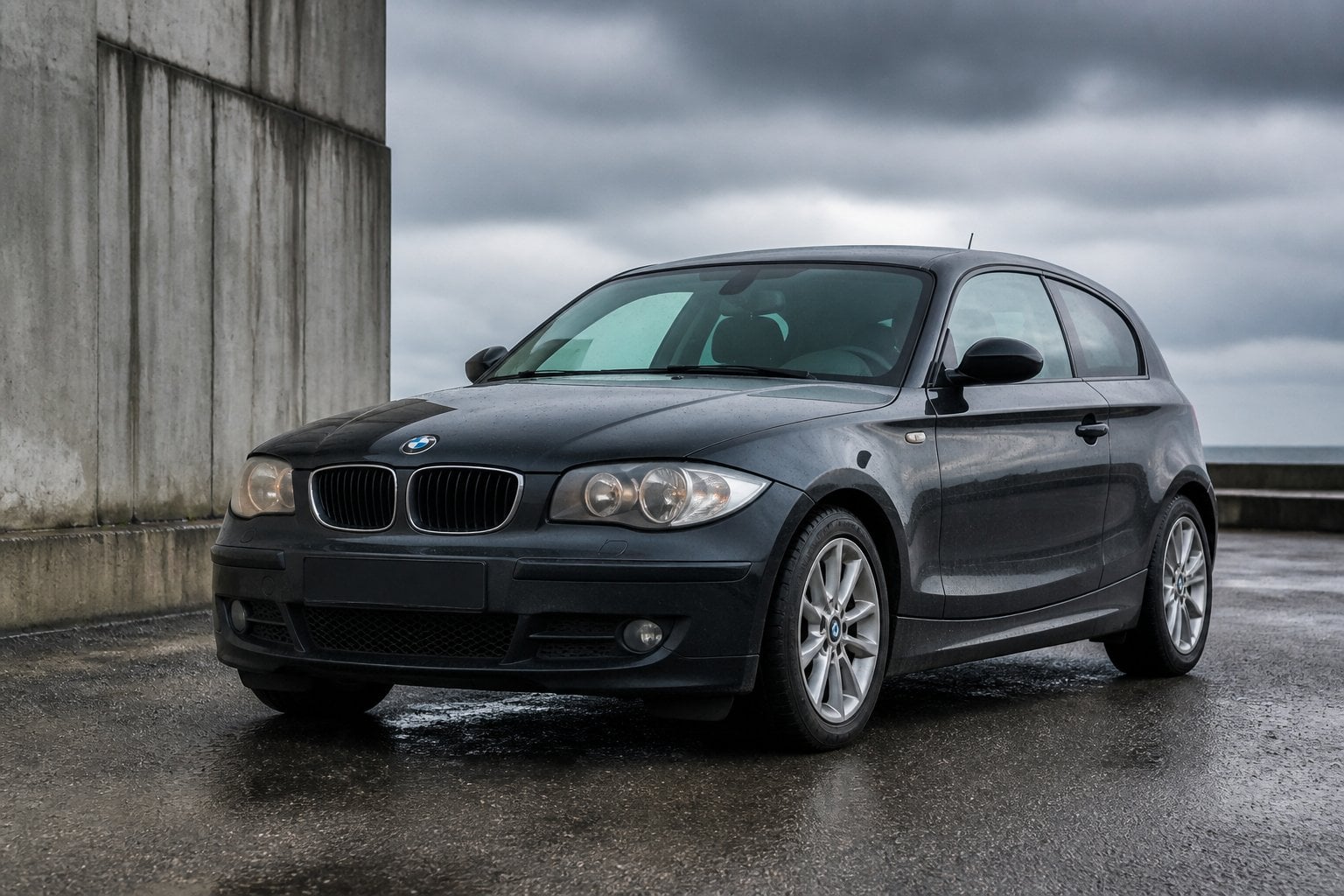 A dark BMW 1 Series parked on a wet industrial road beneath overcast skies.