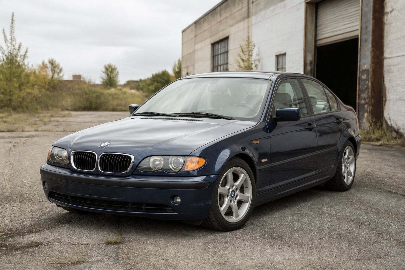A dark blue BMW E46 330i sedan parked outside a weathered industrial garage.