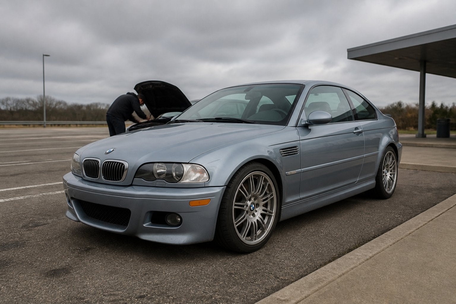 A weathered silver-blue BMW E46 M3 coupe parked at a roadside service area with its hood open.