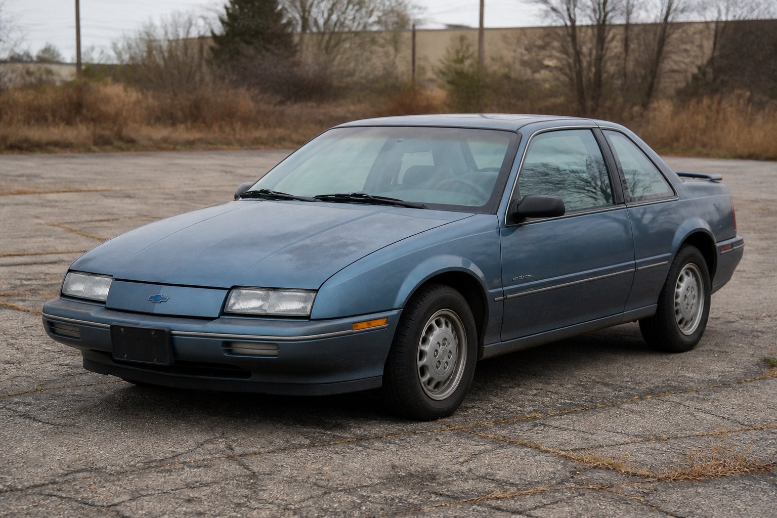 An older Chevrolet Beretta coupe parked in a neglected suburban lot.