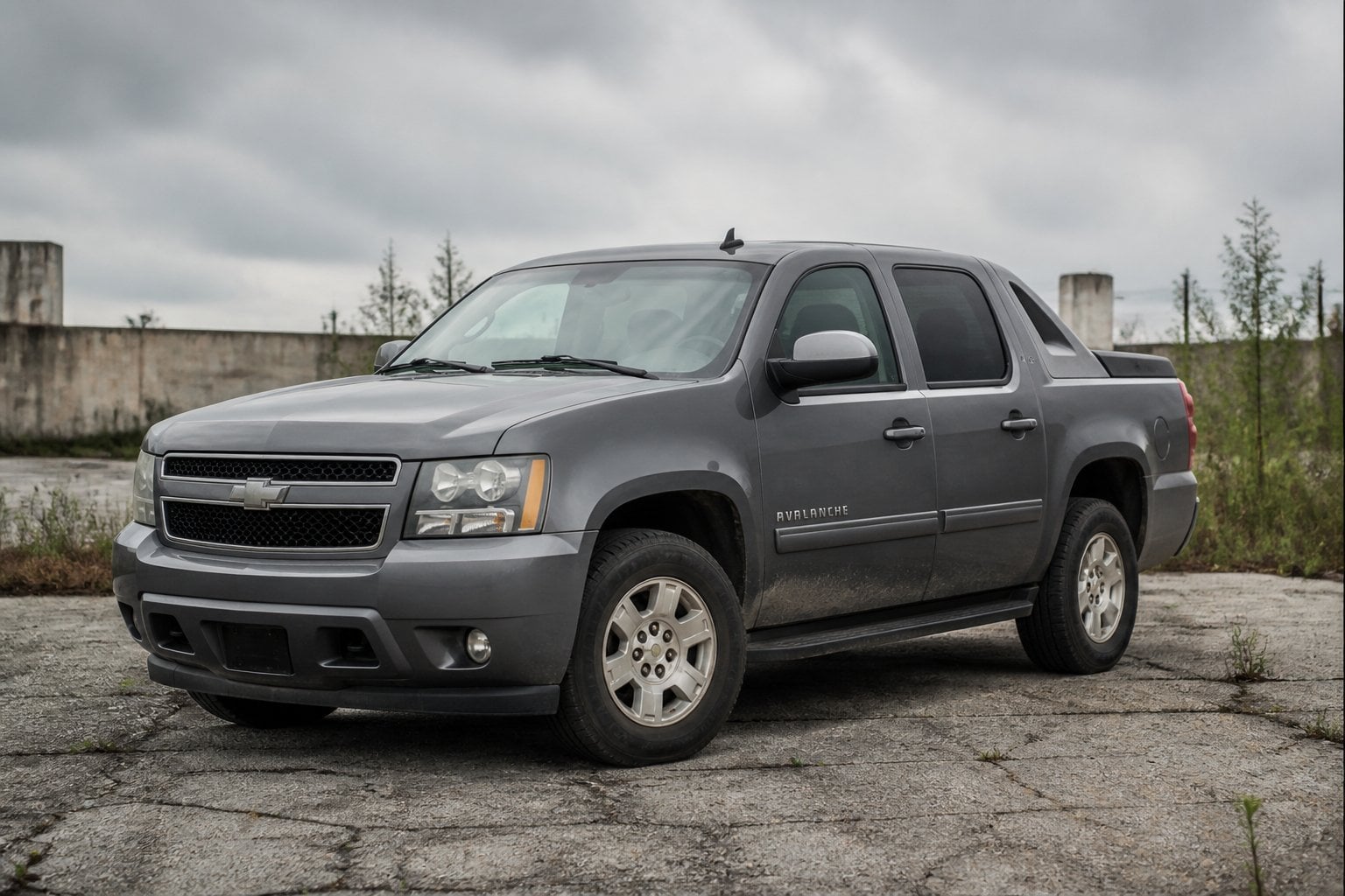 An aging Chevrolet Avalanche pickup truck parked in a neglected industrial lot under an overcast sky.