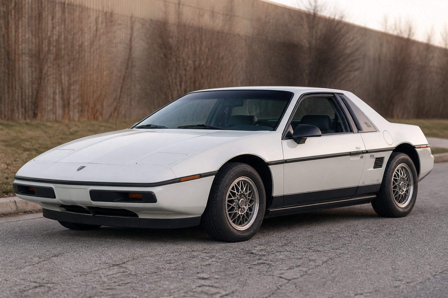 A red 1984 Pontiac Fiero parked on a quiet roadside, viewed from the front three-quarter angle.